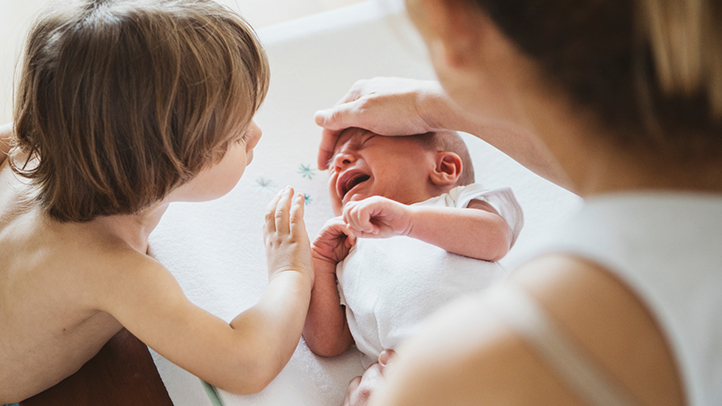 mom checking crying baby's temperature on forehead with older child looking on, croup in babies and toddlers